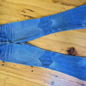 Op de afbeelding: Een paar blauwe denim jeans met een wijde pijp en twee grote, ruitvormige patches op de knieën.
