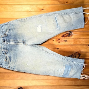 Pode incluir: Calças de ganga azul claro com detalhes desgastados, incluindo remendos e bordas desfiadas, penduradas num cabide de madeira. As calças de ganga estão sobre uma superfície de madeira, mostrando sua aparência desgastada e estilo casual.