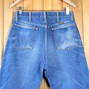 May include: Blue denim jeans with the Wrangler logo on the back pocket. The jeans are hanging on a wooden hanger. The jeans have a classic five-pocket design with a zipper fly and brass rivets.