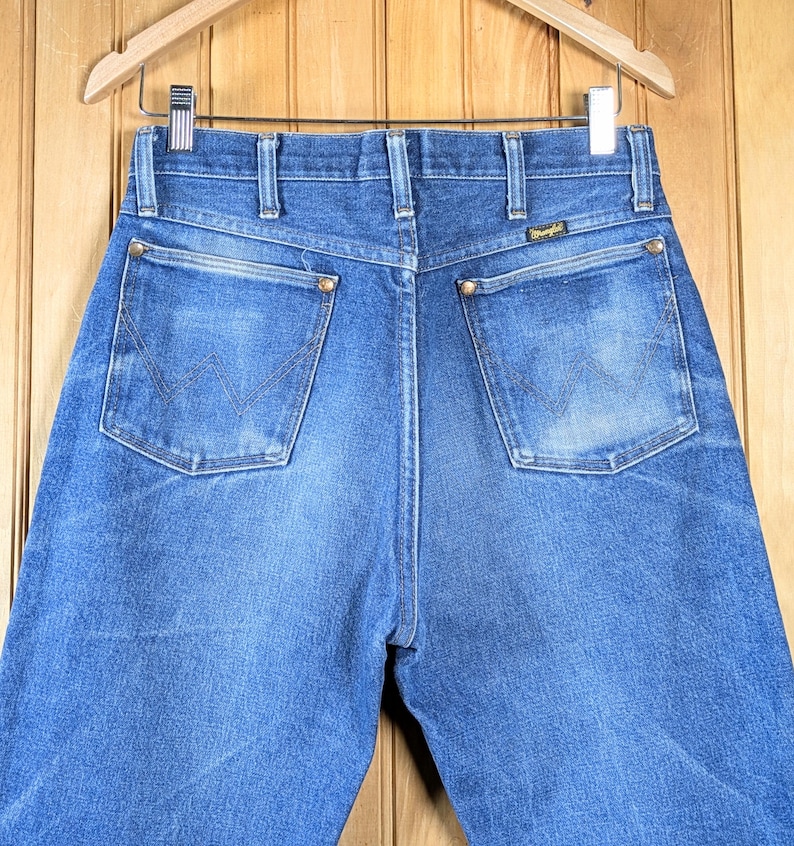 May include: Blue denim jeans with the Wrangler logo on the back pocket. The jeans are hanging on a wooden hanger. The jeans have a classic five-pocket design with a zipper fly and brass rivets.