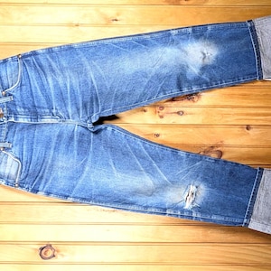 May include: Blue denim jeans with distressed details, displayed on a wooden hanger. The jeans have a faded wash, a ripped knee, and cuffed hems. The background is a wooden surface.