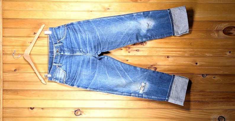 May include: Blue denim jeans with distressed details, displayed on a wooden hanger. The jeans have a faded wash, a ripped knee, and cuffed hems. The background is a wooden surface.