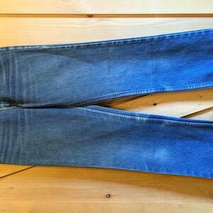 May include: Blue denim jeans with turned-up cuffs, displayed flat on a wooden surface. The jeans feature a classic five-pocket design and a button and zip fastening. The denim has a faded, worn appearance.