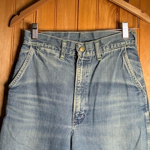 May include: Close-up of blue denim work pants hanging on a wooden hanger. The pants have a button closure, front pockets, and green stitching details. The denim has a faded, worn appearance.