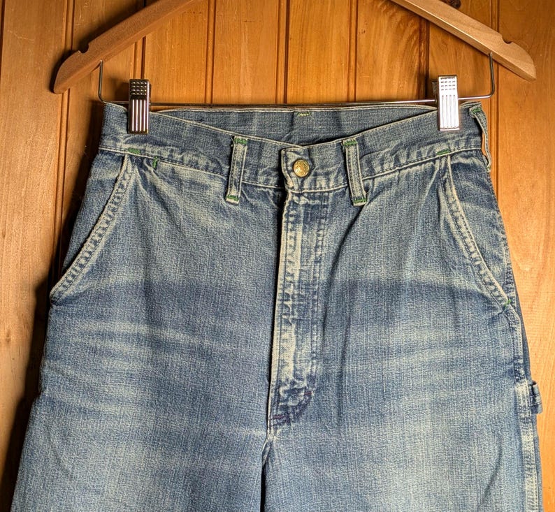 May include: Close-up of blue denim work pants hanging on a wooden hanger. The pants have a button closure, front pockets, and green stitching details. The denim has a faded, worn appearance.