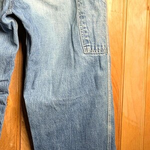 May include: Light blue denim jeans with a classic design, featuring a back pocket and a utility pocket on the leg. The jeans are cuffed at the bottom and hang on a wooden hanger.