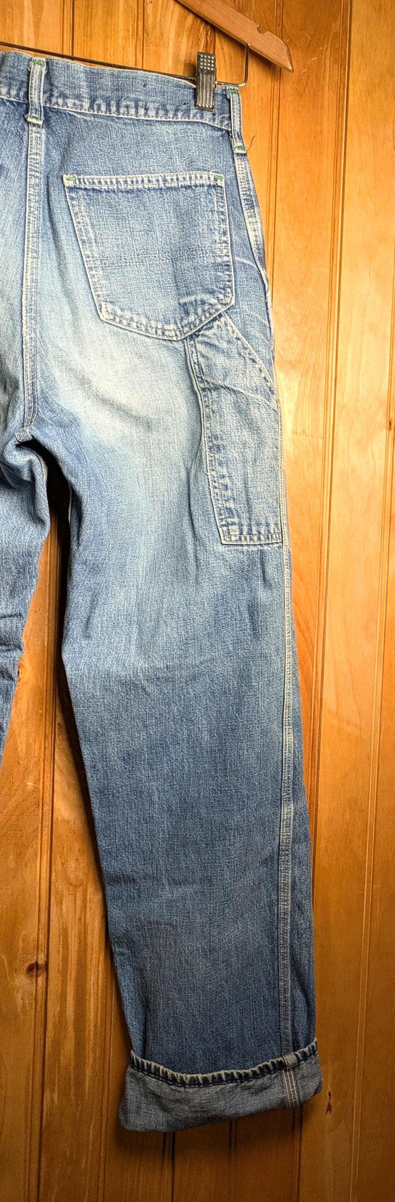 May include: Light blue denim jeans with a classic design, featuring a back pocket and a utility pocket on the leg. The jeans are cuffed at the bottom and hang on a wooden hanger.