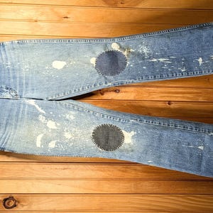 May include: Light blue denim jeans with two dark blue patches on the knees and white paint splatter details. The jeans are hanging on a wooden hanger against a wooden background.
