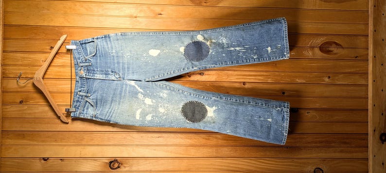 May include: Light blue denim jeans with two dark blue patches on the knees and white paint splatter details. The jeans are hanging on a wooden hanger against a wooden background.