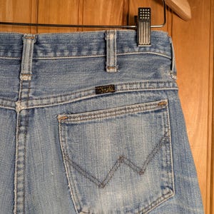 May include: Light blue denim jeans with a classic five-pocket design. The back pocket features a stitched 'W' design. The jeans are hanging on a wooden hanger. The jeans have a small, rectangular brand label.