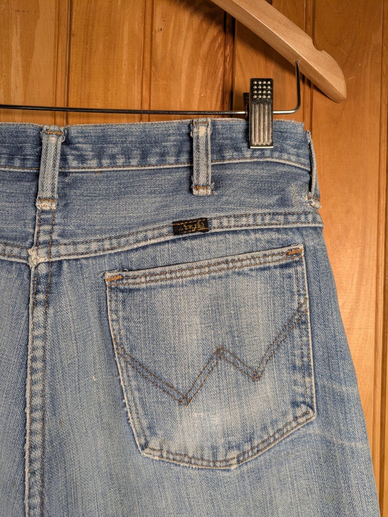 May include: Light blue denim jeans with a classic five-pocket design. The back pocket features a stitched 'W' design. The jeans are hanging on a wooden hanger. The jeans have a small, rectangular brand label.