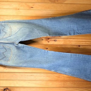 May include: Light blue denim jeans with a flared leg design, hanging on a wooden hanger. The jeans have a faded wash and visible back pockets. The garment is displayed on a wooden surface.