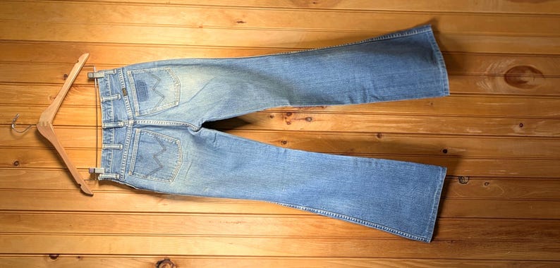 May include: Light blue denim jeans with a flared leg design, hanging on a wooden hanger. The jeans have a faded wash and visible back pockets. The garment is displayed on a wooden surface.