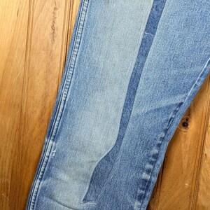 May include: Close-up of light blue denim jeans with a unique, lighter-wash patch design. The jeans show signs of wear and tear, with a faded area and a small tear. The stitching is visible along the seams.