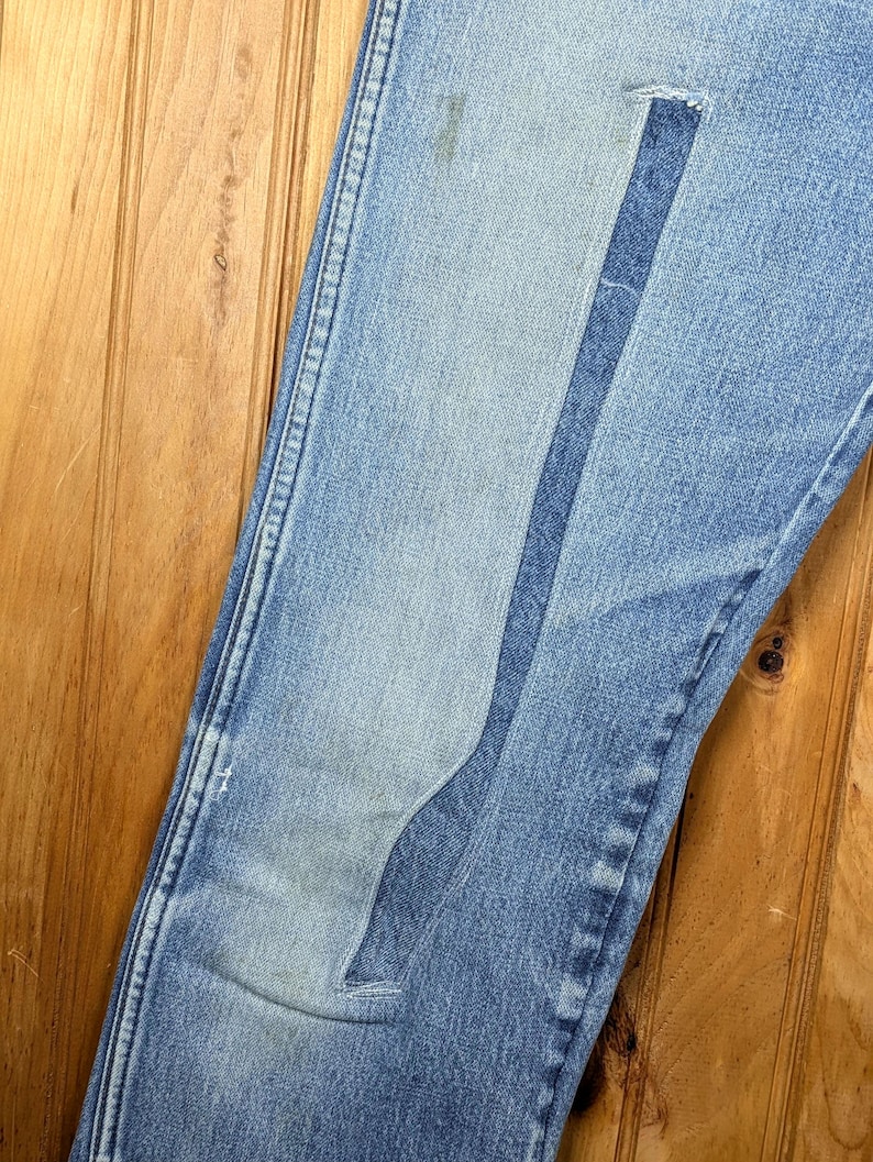 May include: Close-up of light blue denim jeans with a unique, lighter-wash patch design. The jeans show signs of wear and tear, with a faded area and a small tear. The stitching is visible along the seams.
