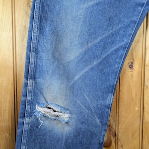 May include: Close-up of blue denim jeans with a distressed, worn look. The jeans have a large tear on the knee, revealing the frayed edges of the fabric. The denim shows fading and wear patterns.