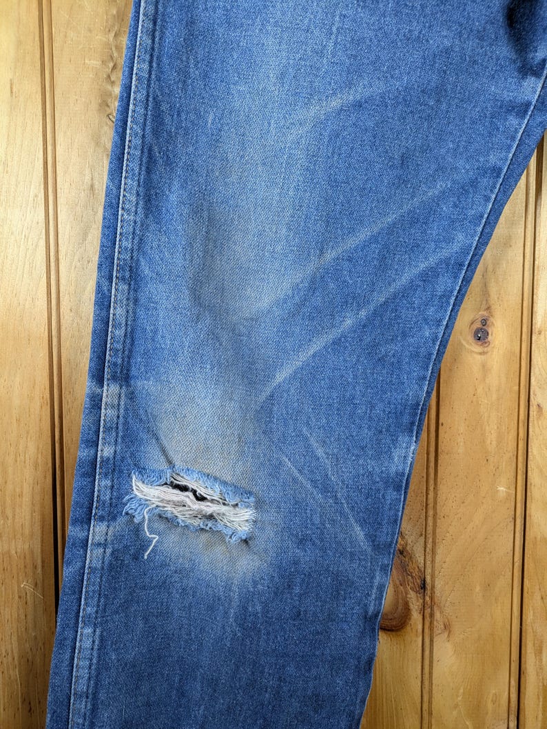 May include: Close-up of blue denim jeans with a distressed, worn look. The jeans have a large tear on the knee, revealing the frayed edges of the fabric. The denim shows fading and wear patterns.