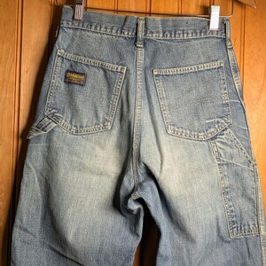May include: Light blue denim shorts with multiple pockets and a faded wash. The shorts feature the "OshKosh" logo on a small patch. The shorts are hanging on a wooden hanger.