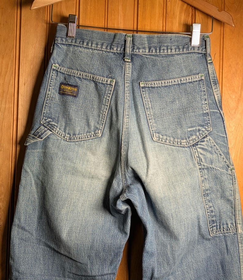 May include: Light blue denim shorts with multiple pockets and a faded wash. The shorts feature the "OshKosh" logo on a small patch. The shorts are hanging on a wooden hanger.
