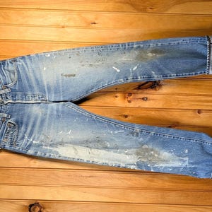 Puede incluir: Vaqueros de mezclilla azul claro con salpicaduras de pintura y puños doblados, colgados de una percha de madera. Los vaqueros están sobre una superficie de madera, mostrando su aspecto desgastado y estilo casual.
