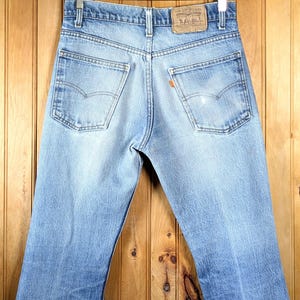 Può includere: Jeans in denim blu chiaro sbiadito, con classico design a cinque tasche e toppa in pelle. I jeans sono appesi a una gruccia di legno. Le tasche posteriori presentano un caratteristico motivo di cucitura.