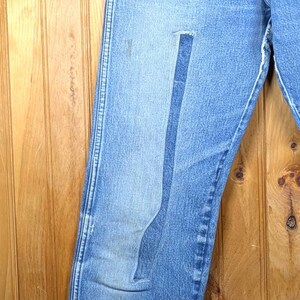May include: Light blue denim jeans with a faded appearance and a patch on the left leg. The jeans are hanging on a wooden hanger against a wooden background. The jeans have a classic five-pocket design.