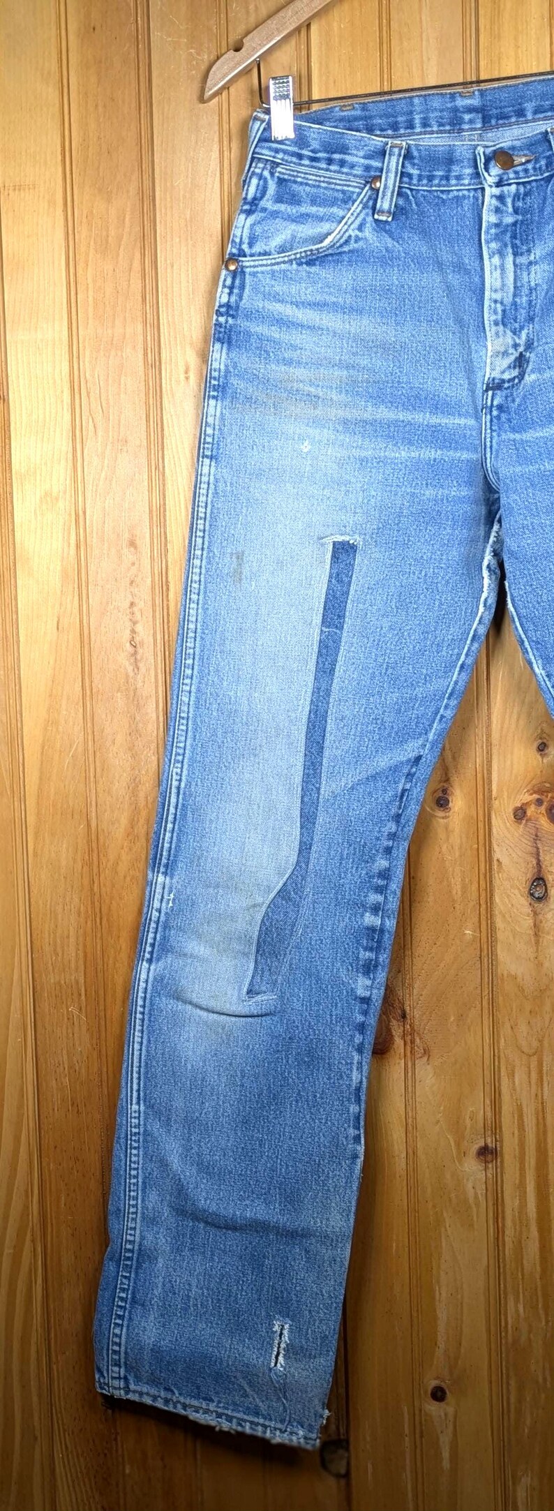 May include: Light blue denim jeans with a faded appearance and a patch on the left leg. The jeans are hanging on a wooden hanger against a wooden background. The jeans have a classic five-pocket design.