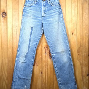 May include: Light blue denim jeans hanging on a wooden hanger. The jeans have a faded appearance with a patch on the left leg and some wear and tear. The jeans are displayed against a wooden background.