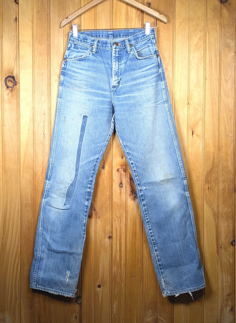 May include: Light blue denim jeans hanging on a wooden hanger. The jeans have a faded appearance with a patch on the left leg and some wear and tear. The jeans are displayed against a wooden background.