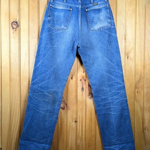 May include: Blue denim jeans with a classic straight leg fit, hanging on a wooden hanger. The jeans have two back pockets and show signs of wear. The background is a wooden wall.