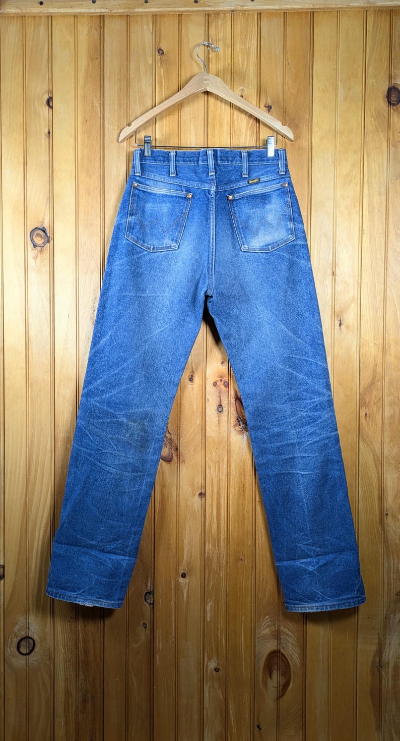 May include: Blue denim jeans with a classic straight leg fit, hanging on a wooden hanger. The jeans have two back pockets and show signs of wear. The background is a wooden wall.