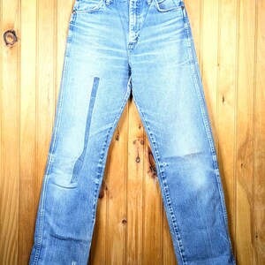 May include: Light blue denim jeans hanging on a wooden hanger. The jeans have a high waist, a straight leg, and a faded wash. The jeans are displayed against a wooden background.