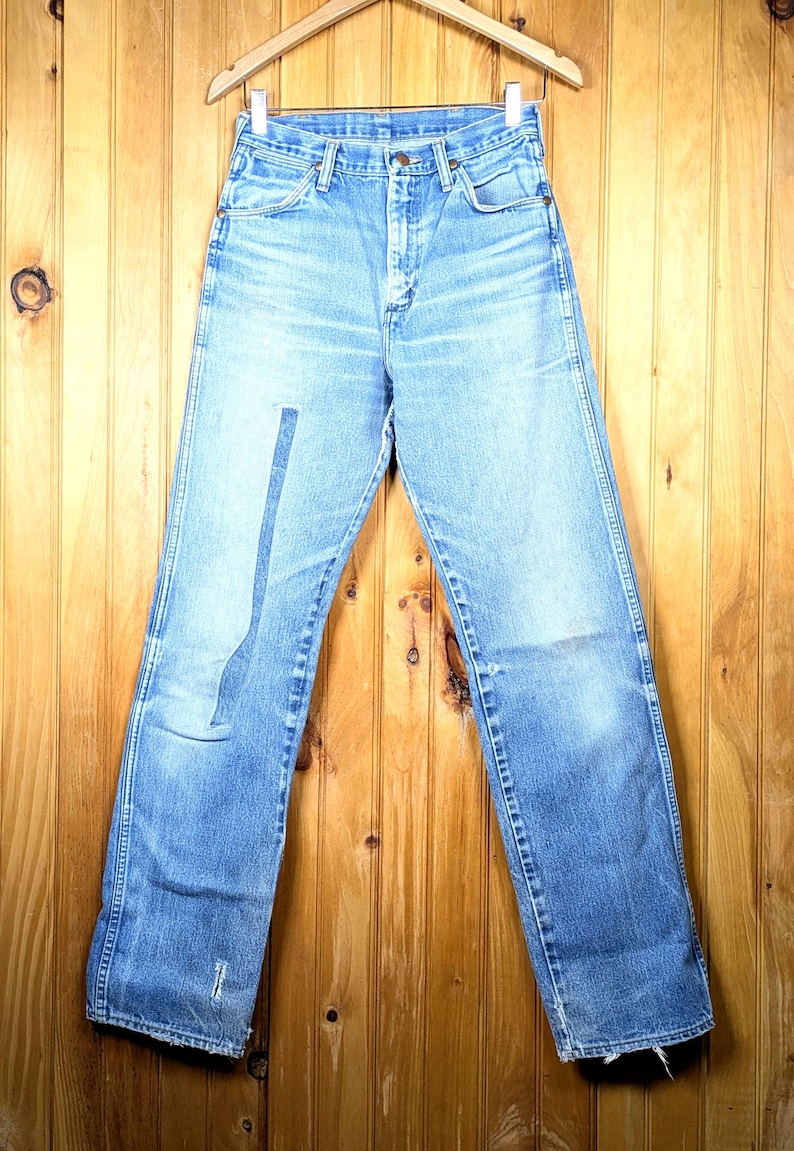 May include: Light blue denim jeans hanging on a wooden hanger. The jeans have a high waist, a straight leg, and a faded wash. The jeans are displayed against a wooden background.