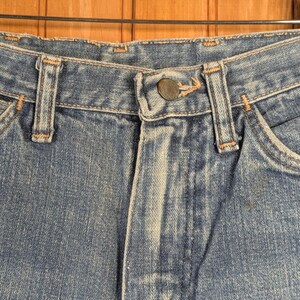 May include: Close-up of blue denim jeans, showing the button closure, belt loops, and pocket details. The jeans have a classic, worn look with visible stitching and a faded appearance, suggesting they are vintage or well-loved.