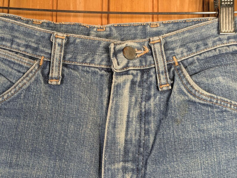 May include: Close-up of blue denim jeans, showing the button closure, belt loops, and pocket details. The jeans have a classic, worn look with visible stitching and a faded appearance, suggesting they are vintage or well-loved.