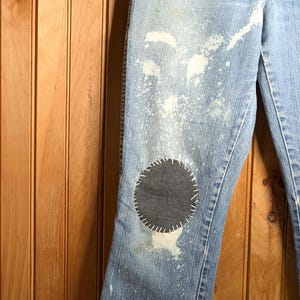 May include: Light blue denim jeans with a distressed look, featuring white paint splatters and a large, dark gray circular patch on the knee. The jeans are hanging on a wooden hanger.