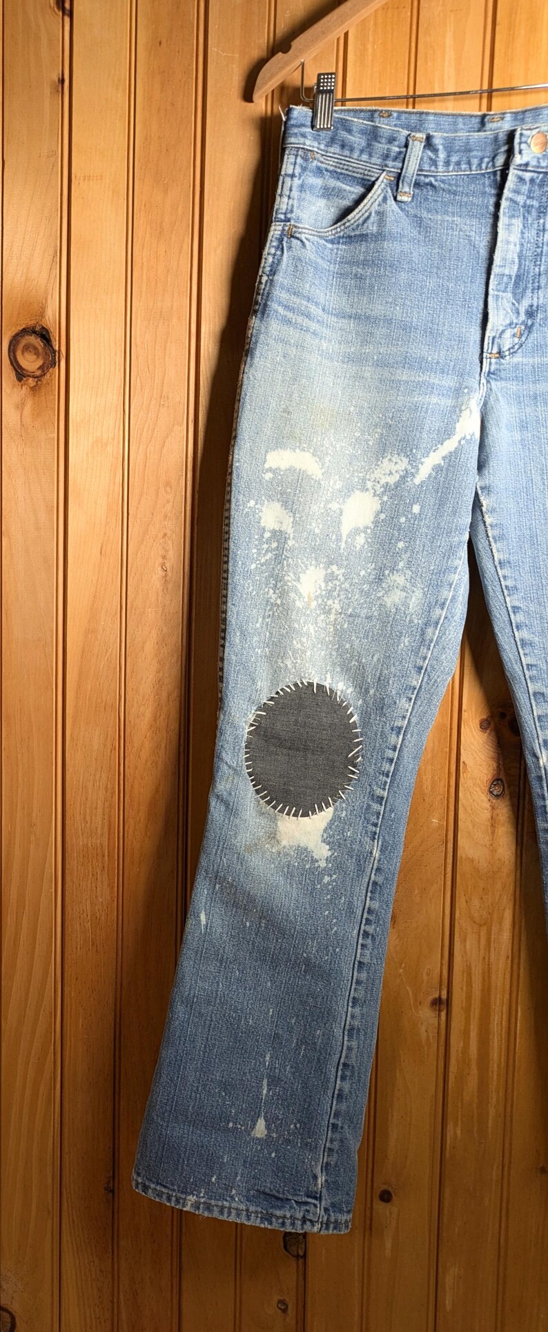 May include: Light blue denim jeans with a distressed look, featuring white paint splatters and a large, dark gray circular patch on the knee. The jeans are hanging on a wooden hanger.