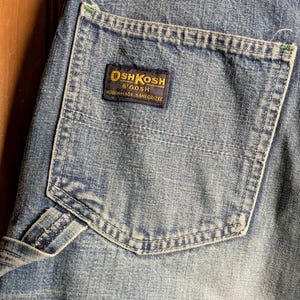 May include: Close-up of light blue denim fabric with a pocket. The pocket features a rectangular label with the text "OSH KOSH B'GOSH UNION MADE SANFORIZED" in gold and navy blue. The denim has visible stitching and a worn appearance.