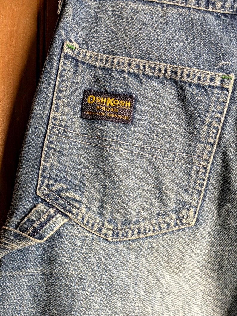 May include: Close-up of light blue denim fabric with a pocket. The pocket features a rectangular label with the text "OSH KOSH B'GOSH UNION MADE SANFORIZED" in gold and navy blue. The denim has visible stitching and a worn appearance.
