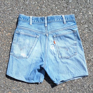 May include: Light blue denim shorts featuring a high-waisted design with belt loops, two back pockets with stitched arcuate detailing, and a small orange tab on the right pocket. The fabric shows signs of wear with light fading and fraying.