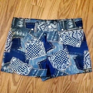 May include: Blue denim shorts with a patchwork design featuring various shades of blue and white. The shorts have a high waist and a relaxed fit.