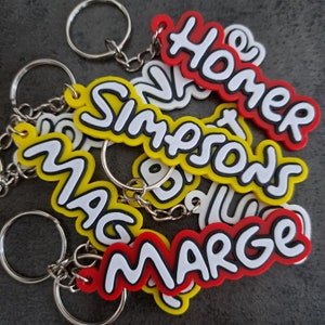 Może przedstawiać: Zestaw sześciu breloków z nazwami "Homer", "Marge", "Bart", "Lisa", "Maggie" i "Simpsonowie" w kreskówkowym stylu. Breloki są wykonane z kolorowego plastiku i mają srebrne kółka.