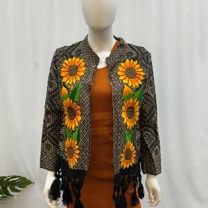 Peut inclure: Veste brodée noire et marron avec un motif de tournesols. La veste présente des tournesols jaunes avec des feuilles vertes et des franges noires. Elle est présentée sur un mannequin portant une robe orange. Vêtement unique.