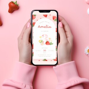 May include: A pink smartphone displaying a digital invitation for Amelia's 3rd birthday party. The invitation features a strawberry and flower design with the text "Please join us as Amelia is turning 3". The date is September 30th, 2023 at 4:00pm.
