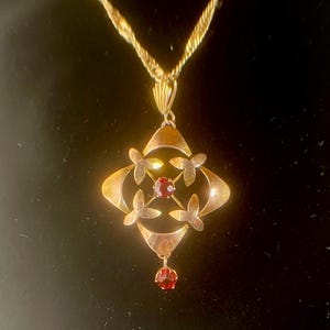 May include: Gold-toned pendant necklace featuring a geometric design with four floral accents and a central red gemstone. A smaller red gemstone dangles below. The necklace has a twisted rope chain.