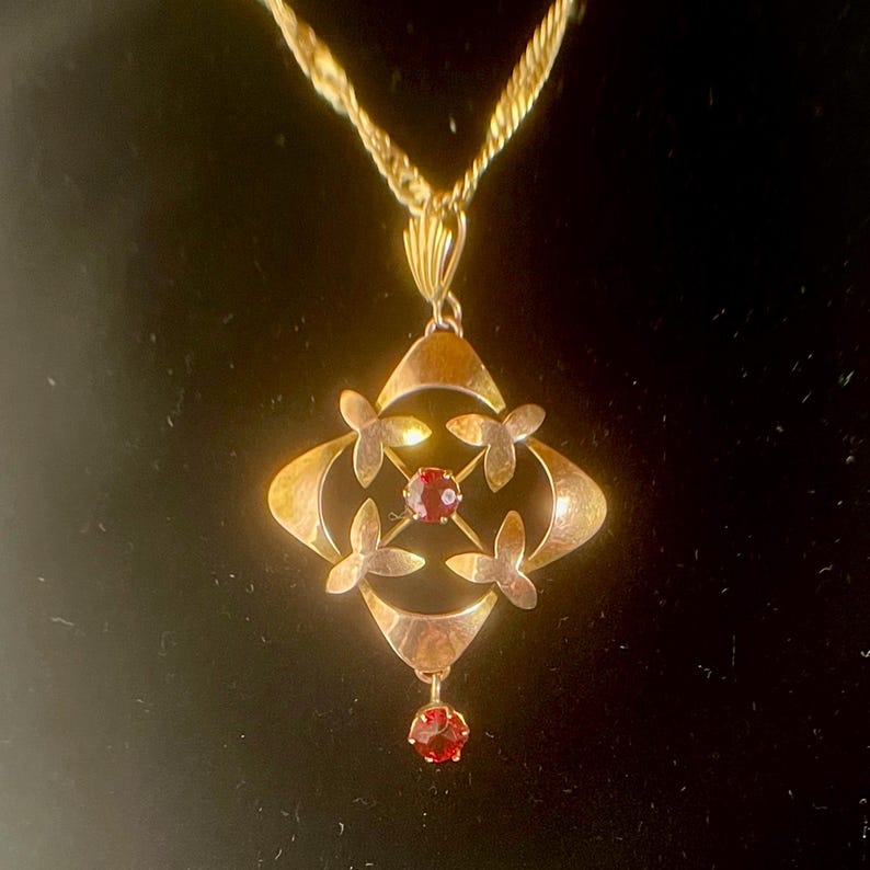 May include: Gold-toned pendant necklace featuring a geometric design with four floral accents and a central red gemstone. A smaller red gemstone dangles below. The necklace has a twisted rope chain.