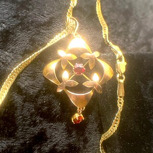 May include: A gold-toned pendant necklace with a diamond-shaped centerpiece. The pendant features a central red gemstone, surrounded by four stylized floral elements. A smaller red gemstone hangs below. The necklace chain is also gold-toned.