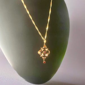 May include: A gold-colored pendant necklace with a twisted chain. The pendant has a four-leaf clover design with small red gemstones in the center and at the bottom. The necklace is displayed on a dark green surface.