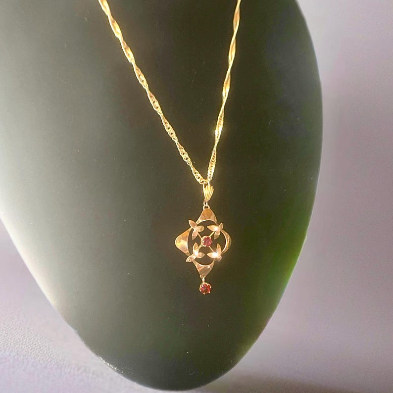 May include: A gold-colored pendant necklace with a twisted chain. The pendant has a four-leaf clover design with small red gemstones in the center and at the bottom. The necklace is displayed on a dark green surface.