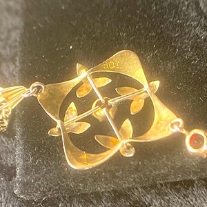 May include: A gold-toned brooch with a geometric design featuring leaf-like cutouts. The brooch has a pin closure and a small red gemstone pendant. The metal is stamped with "9ct".
